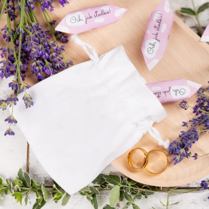 Elegancki biały woreczek satynowy obok złotych obrączek i kwiatów lawendy.